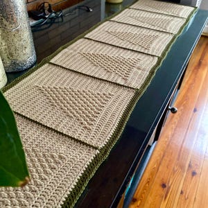 Peut inclure: Chemin de table beige crocheté à la main avec une bordure verte. Le chemin de table présente une série de motifs carrés texturés. Le chemin de table est présenté sur une surface sombre, mettant en valeur les détails du travail artisanal.