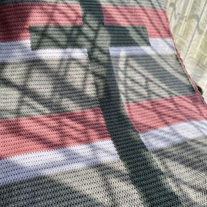 May include: A handmade crochet blanket featuring a striped pattern in shades of gray, white, and dusty rose. The blanket's texture is visible, with a close-up view highlighting the detailed stitching. The design includes a dark green stripe.