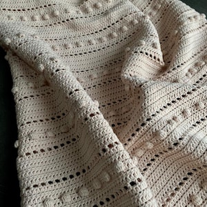 Pode incluir: Uma manta de crochê dobrada, de cor creme. A manta apresenta um padrão texturizado com fileiras de pequenas bolinhas em relevo e pontos vazados. A cor suave e neutra e o design intrincado sugerem um item feito à mão.