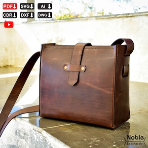 May include: A brown leather shoulder bag with a rectangular shape. The bag features a flap closure with a strap and brass-coloured hardware. The bag has a long, adjustable shoulder strap. The words "Noble Leather Art" are in the bottom right corner.