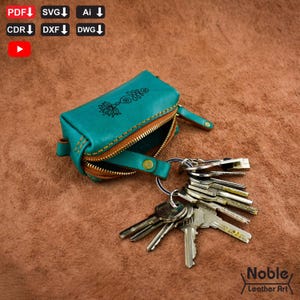 May include: Teal leather key pouch with a brown zipper and a decorative floral design. The pouch is open, revealing the interior, and is attached to a keyring with multiple keys. The pouch is approximately 4 inches long.