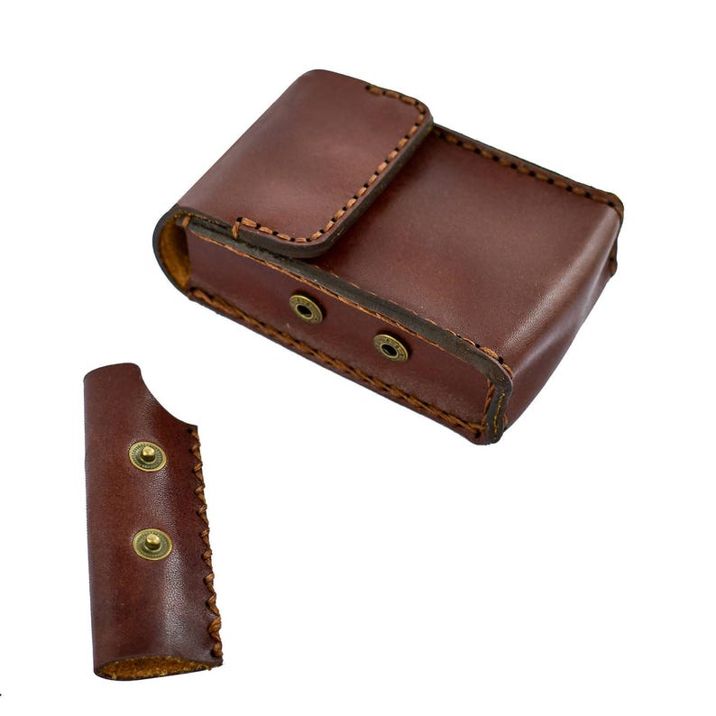 May include: A brown leather case with a flap closure and two brass-colored snap closures. The case is rectangular and appears to be hand-stitched. A separate, smaller leather piece with two snaps is also visible.