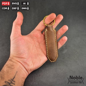 Puede incluir: Un llavero de cuero marrón con un llavero dorado. El llavero es rectangular con un borde cosido con un hilo marrón más claro. El texto "Noble Leather Art" está en la esquina inferior derecha.