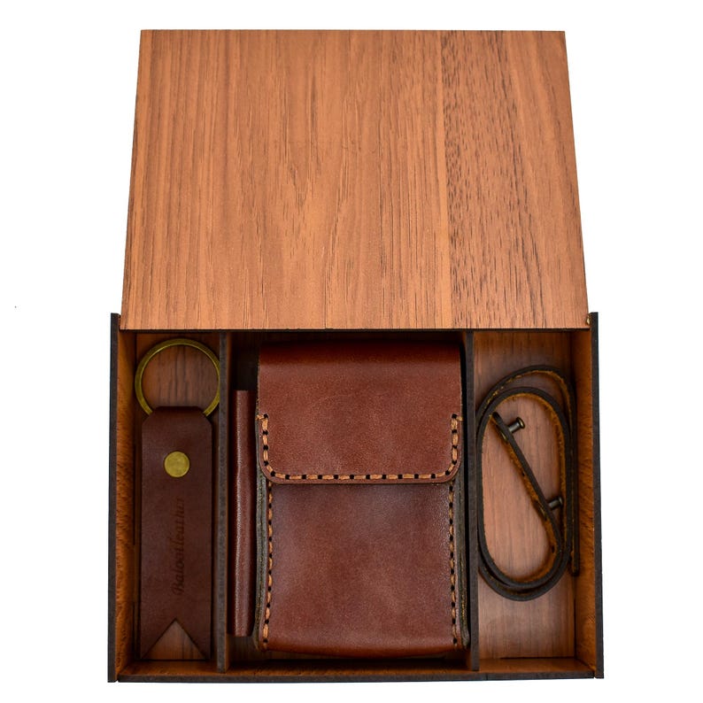 May include: A wooden gift box with a hinged lid, containing a brown leather wallet, a keychain, a pen, and a leather neck strap. The leather items have contrasting stitching. The box is made of light brown wood.