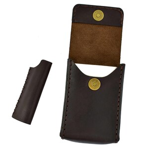 May include: A dark brown leather business card holder with a magnetic snap closure. The holder is open, revealing a light brown interior. A separate, matching leather piece is positioned to the left.