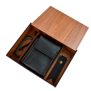 May include: A wooden box with a hinged lid, containing a black leather wallet, a black leather keychain, and a black carabiner clip. The box and lid are made of wood with a natural finish.