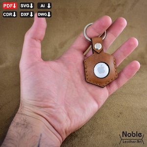 May include: A tan leather AirTag holder with a silver key ring. The holder is hexagonal with a stitched border and a circular opening for the AirTag. The Apple logo is visible. The holder is held in a hand.