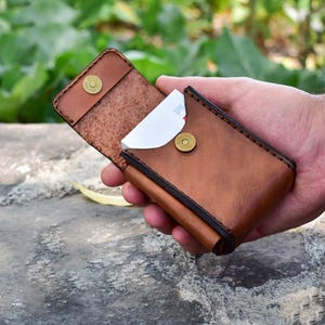 May include: A brown leather card holder with a snap closure, open to reveal white cards. The wallet is held in a hand, showcasing the craftsmanship and design. The leather has a rich, textured appearance.