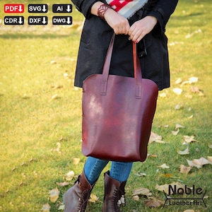 May include: A burgundy leather tote bag with long handles is held by a person. The bag has a smooth, polished finish and visible stitching along the sides. The words "Noble Leather Art" are in the bottom right corner.