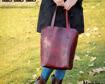 Puede incluir: Un bolso tote de cuero burdeos con asas largas es sostenido por una persona. El bolso tiene un acabado liso y pulido y costuras visibles en los laterales. Las palabras "Noble Leather Art" están en la esquina inferior derecha.