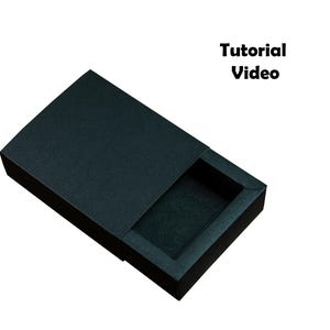 May include: A black, textured, rectangular box with a sliding lid, partially open to reveal a dark interior. The text "Tutorial Video" is displayed in bold, black font.