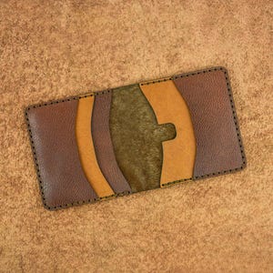 May include: A handmade leather card wallet in shades of brown, tan, and olive green. The wallet features a unique design with curved accents and visible stitching around the edges. The wallet is rectangular and appears to be empty.