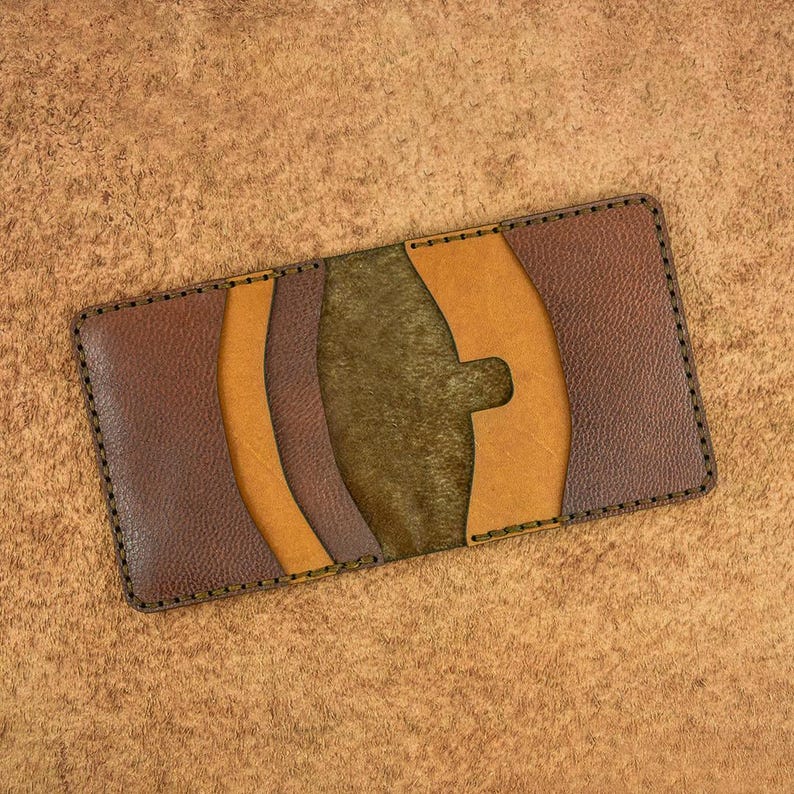 May include: A handmade leather card wallet in shades of brown, tan, and olive green. The wallet features a unique design with curved accents and visible stitching around the edges. The wallet is rectangular and appears to be empty.