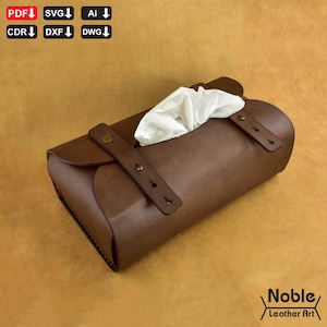 Könnte beinhalten: Eine braune Leder-Taschentuchbox mit rechteckiger Form und Klappenverschluss. Weiße Taschentücher sind oben sichtbar. Der Bezug hat zwei Lederriemen mit messingfarbenen Nieten. Der Text "Noble Leather Art" befindet sich unten rechts.