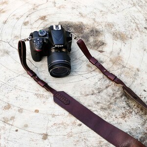 K&ouml;nnte beinhalten: Eine schwarze Nikon DSLR-Kamera mit Objektiv und einem dunkelbraunen Kameragurt aus Leder. Der Gurt ist gen&auml;ht und hat Metallverschl&uuml;sse. Die Kamera und der Gurt liegen auf einer hellen, strukturierten Oberfl&auml;che.