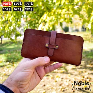 Könnte beinhalten: Eine braune Ledergeldbörse mit Riemenverschluss und messingfarbenen Nieten. Die Geldbörse wird in einer Hand vor einem verschwommenen Hintergrund aus grünem Laub gehalten. Die Worte "Noble Leather Art" befinden sich in der unteren rechten Ecke.