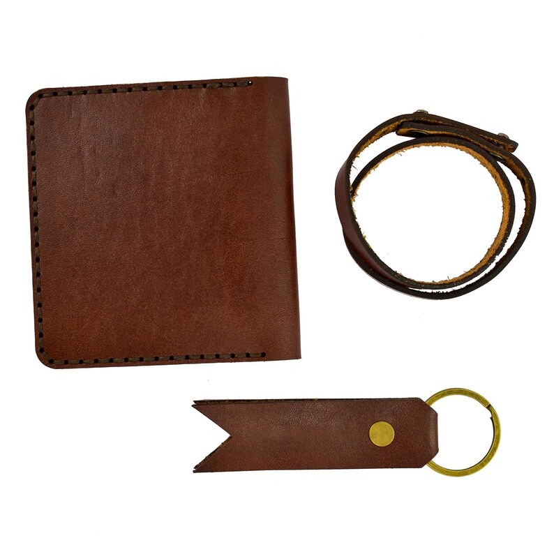 May include: A brown leather wallet, bracelet, and keychain set. The wallet is rectangular with stitched edges. The bracelet is a wrap-around style. The keychain has a gold-colored ring and a decorative end.