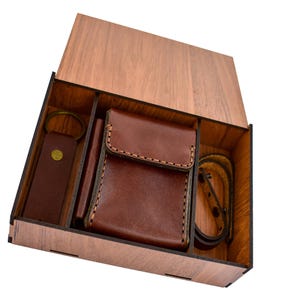 May include: A wooden box with a hinged lid, containing a brown leather wallet, key ring, and bracelet. The box and accessories are made of wood and leather. The wallet has a stitched border.