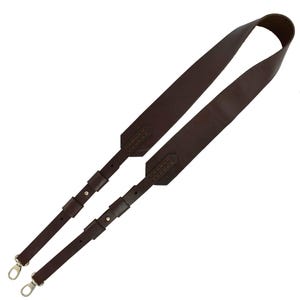 K&ouml;nnte beinhalten: Ein dunkelbrauner Leder-Schulterriemen mit silberfarbenen Metallverschl&uuml;ssen. Der Riemen hat einen breiten, gepolsterten Bereich f&uuml;r die Schulter und schmalere, verstellbare Riemen. Das Leder ist f&uuml;r die Haltbarkeit gen&auml;ht.