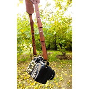 K&ouml;nnte beinhalten: Eine schwarze DSLR-Kamera mit einem braunen Leder-Kameragurt, der an einem Baum h&auml;ngt. Der Gurt ist verstellbar und hat dekorative N&auml;hte. Der Hintergrund zeigt verschwommene gr&uuml;ne B&auml;ume und gelbe Bl&auml;tter.