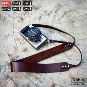 Op de afbeelding: Een zilveren digitale camera met een zwarte lensdop en een bruine leren riem. De riem is met metalen sluitingen aan de camera bevestigd. De camera ligt op een getextureerd oppervlak. De tekst "Noble Leather Art" staat rechtsonder.