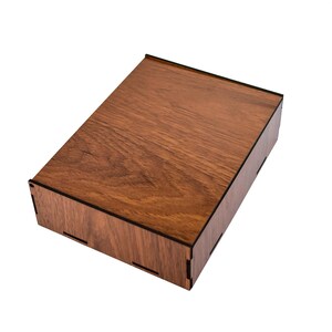May include: A rectangular wooden box with a lid. The box has a dark brown wood grain pattern and black trim. The box is closed and appears to be empty. The box is made of wood.