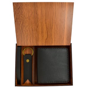 May include: A wooden gift box with a hinged lid, revealing a black leather wallet, a matching key fob with a gold-colored ring, and a black leather belt. The box has a natural wood grain finish.