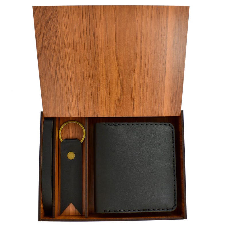 May include: A wooden gift box with a hinged lid, revealing a black leather wallet, a matching key fob with a gold-colored ring, and a black leather belt. The box has a natural wood grain finish.