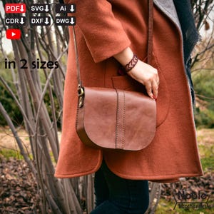 May include: A brown leather crossbody bag with a curved flap and stitched details. The bag is worn over a rust-colored coat. The image includes text that says "in 2 sizes" and "Noble leather art".