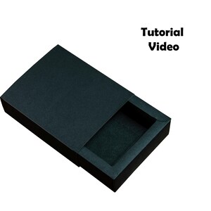 May include: A black, rectangular box with a sliding lid, partially open to reveal a dark interior. The box has a textured surface. The words "Tutorial Video" are printed in black.