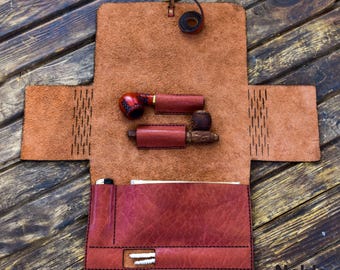 May include: Open leather tobacco pipe case in shades of brown and red. The case holds two pipes, each in a protective leather sleeve. The case is open, revealing a pocket and a loop closure. The logo "Noble Leather Art" is visible.