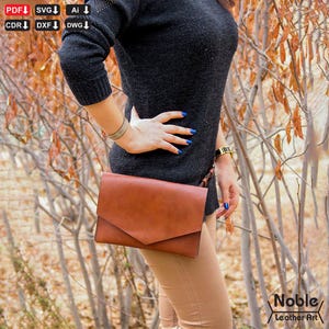 Pode incluir: Uma clutch de couro castanho com fecho de aba. A clutch é segurada contra uma pessoa que usa um suéter preto e calças bege. A imagem inclui o texto "Noble Leather Art".