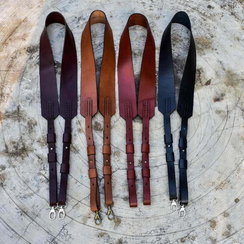 K&ouml;nnte beinhalten: F&uuml;nf Kameragurte aus Leder in verschiedenen Farben: Dunkelbraun, Mittelbraun, Rotbraun, Marineblau und Schwarz. Jeder Gurt ist in der L&auml;nge verstellbar und hat Metall-Drehhaken. Sie werden auf einer hellen, strukturierten Oberfl&auml;che pr&auml;sentiert.