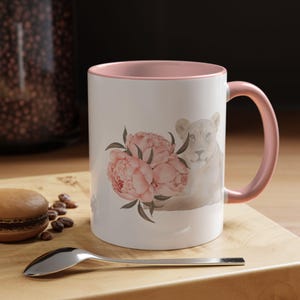 May include: White ceramic mug with a pink interior and handle. The mug features a watercolor design of pink peonies and a lioness. A silver spoon and a macaron are in the foreground.