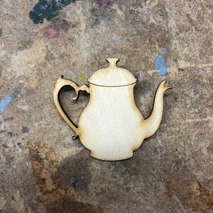 May include: A wooden cutout of a teapot with a simple design. The teapot is light brown and has a rounded body with a spout and a handle.