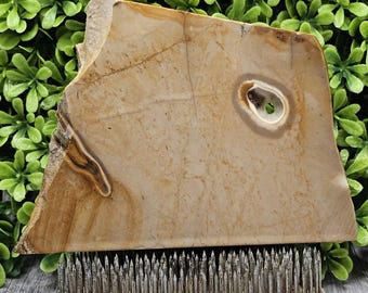 May include: A large, irregularly shaped stone slab with a natural, beige and brown color palette. The stone has a hole and is displayed on a silver metal flower frog, a tool used in floral arrangements. The background features green foliage.