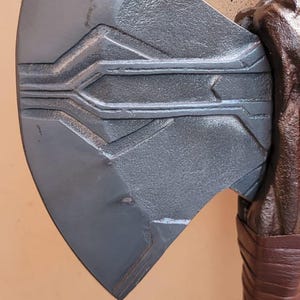 May include: Close-up of a gray metal axe head with etched geometric designs. The axe head is attached to a brown wooden handle wrapped with brown leather. The text "ThelronCreations" is visible.