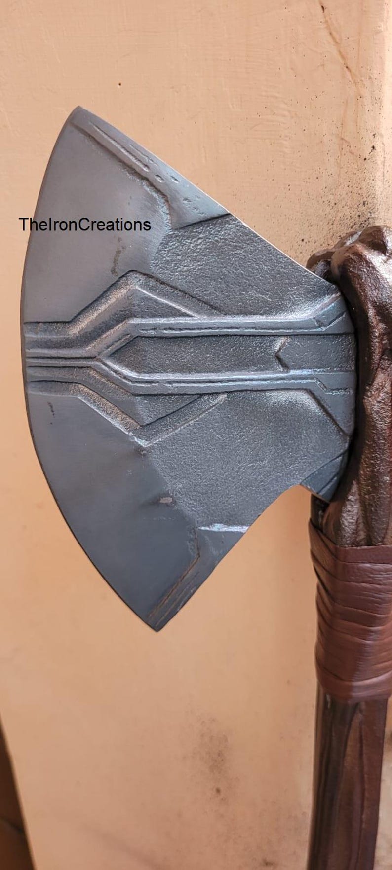 May include: Close-up of a gray metal axe head with etched geometric designs. The axe head is attached to a brown wooden handle wrapped with brown leather. The text "ThelronCreations" is visible.