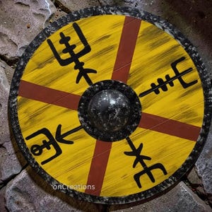 May include: A circular wooden shield featuring a yellow base with a dark red cross pattern. Four black runic symbols are painted in the quadrants. The shield has a central metallic dome and a textured black border with white speckling.