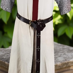 May include: Cream-colored medieval tunic with a large burgundy cross on the chest. The sleeves are chainmail, and a black leather belt with a brass buckle cinches the waist. The tunic is split in the front.