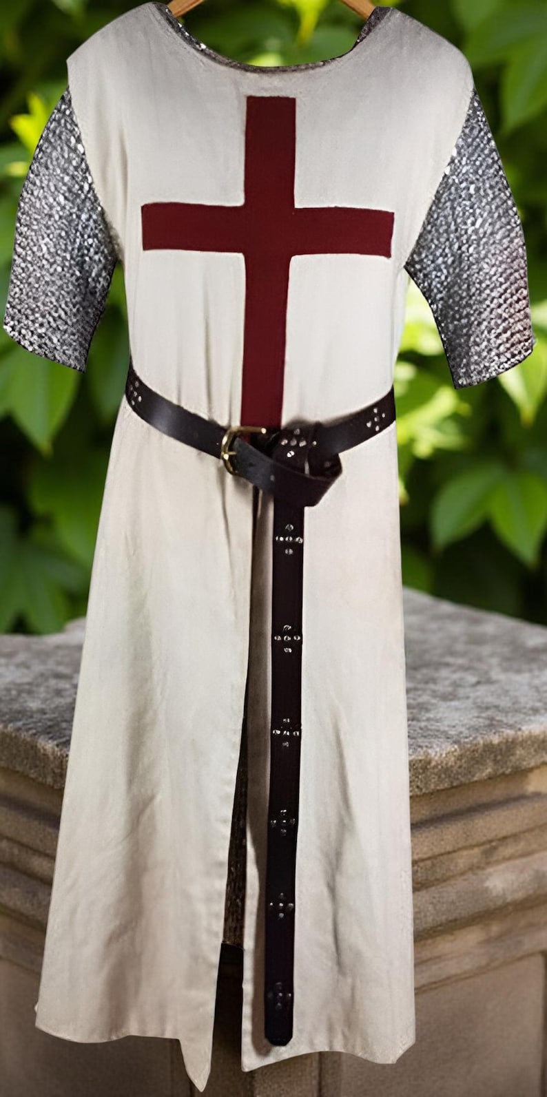 May include: Cream-colored medieval tunic with a large burgundy cross on the chest. The sleeves are chainmail, and a black leather belt with a brass buckle cinches the waist. The tunic is split in the front.