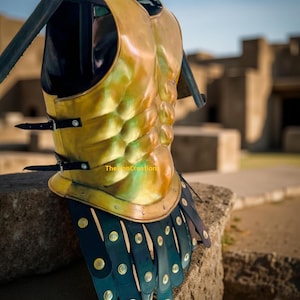 May include: A golden breastplate with black leather straps and accents. The armor has a ribbed design on the front and is secured with buckles. The bottom edge features layered, dark blue leather plates with gold rivets. The text "TheLionCreation" is visible.