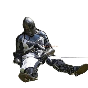 May include: A person in full medieval armor, including a helmet, chest plate, and leg protection. The armor is primarily black and silver, with white accents. The person is seated on the ground, suggesting a historical reenactment or costume.