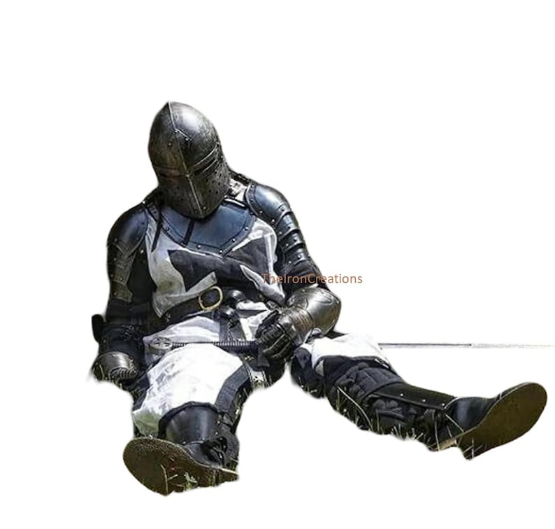 May include: A person in full medieval armor, including a helmet, chest plate, and leg protection. The armor is primarily black and silver, with white accents. The person is seated on the ground, suggesting a historical reenactment or costume.