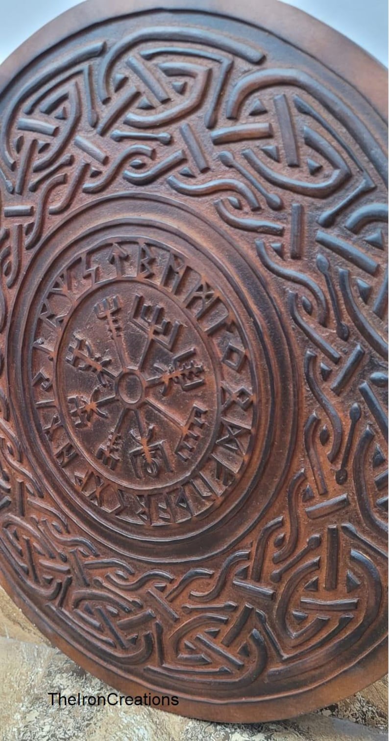 May include: A round, brown, wooden shield with intricate carved Viking knotwork and a central compass design. The shield features a weathered, rustic appearance, with the text "TheIronCreations" at the bottom.
