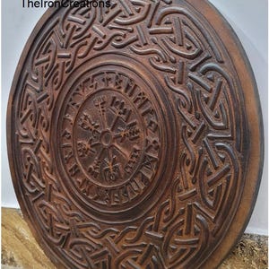 May include: A circular, brown wooden shield with detailed carved knotwork and a central compass design. The shield has the text "TheIronCreations" and is likely a decorative item or wall hanging.