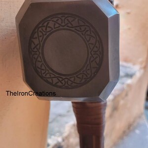 May include: A dark gray metal hammer with a geometric design on the head and a brown handle. The text "ThelronCreations" is visible on the side of the hammer.