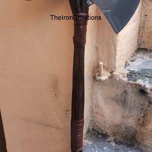 May include: A large, gray and brown axe with a wooden handle and a detailed axe head. The handle is wrapped with brown leather. The text "ThelronCreations" is visible on the axe.