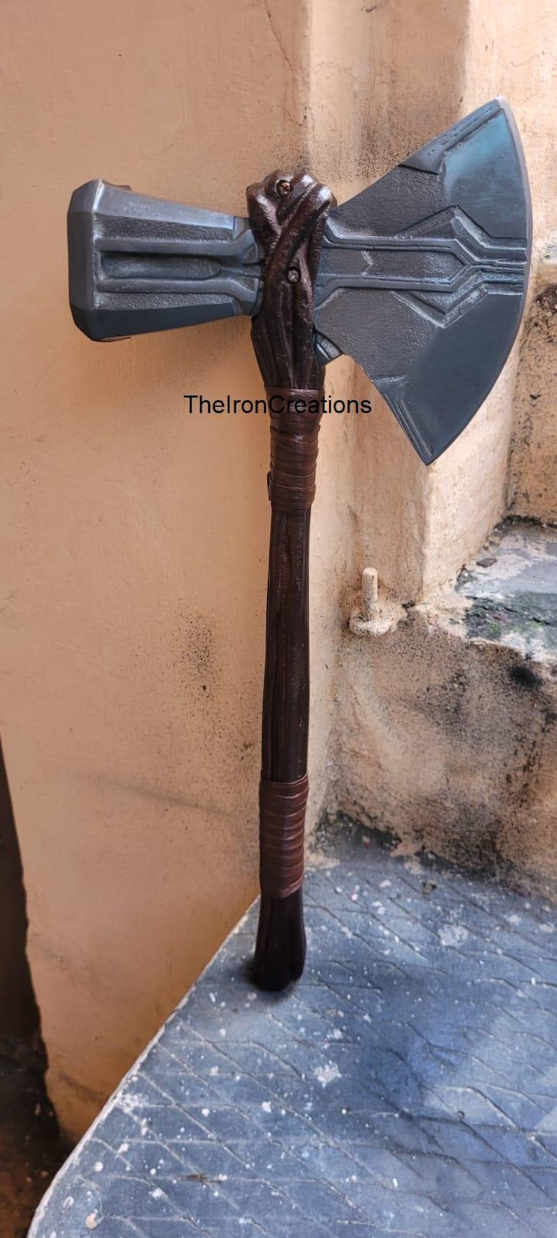 May include: A large, gray and brown axe with a wooden handle and a detailed axe head. The handle is wrapped with brown leather. The text "ThelronCreations" is visible on the axe.