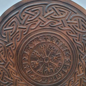May include: A round, brown, wooden item with intricate Celtic knotwork and a central compass design. The item features a weathered, textured appearance and the text "TheIronCreations" at the bottom. The design is reminiscent of Viking or Norse art.